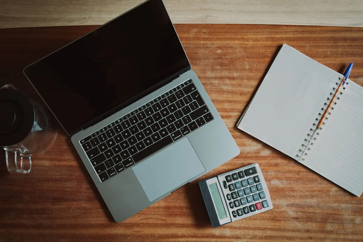 Laptop, calculator and open notepad on a wooden desk, seen from above